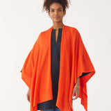 Female wearing a bright orange wrap over blue jeans and top smiles in front of white wall