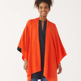 Female wearing a bright orange wrap over blue jeans and top smiles in front of white wall