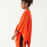 Female wearing a bright orange wrap over blue jeans and top smiles in front of white wall