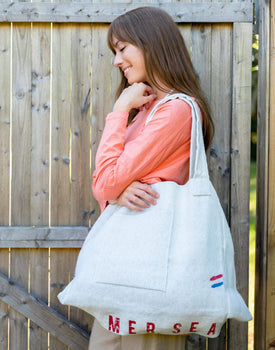 female wearing large white MERSEA labeled tote bag over one shoulder standing outside near a fence 