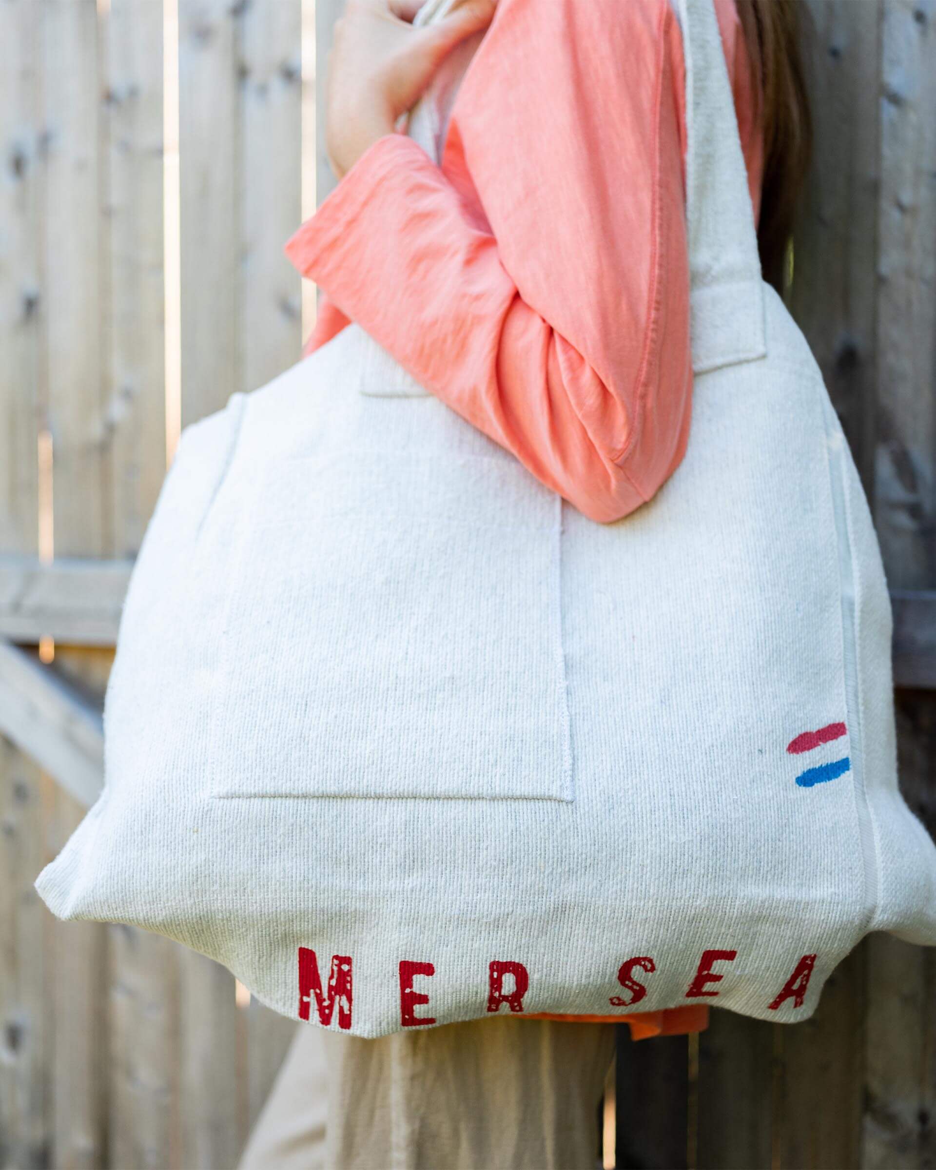 close up of female wearing large white MERSEA labeled tote bag standing outside near a fence 