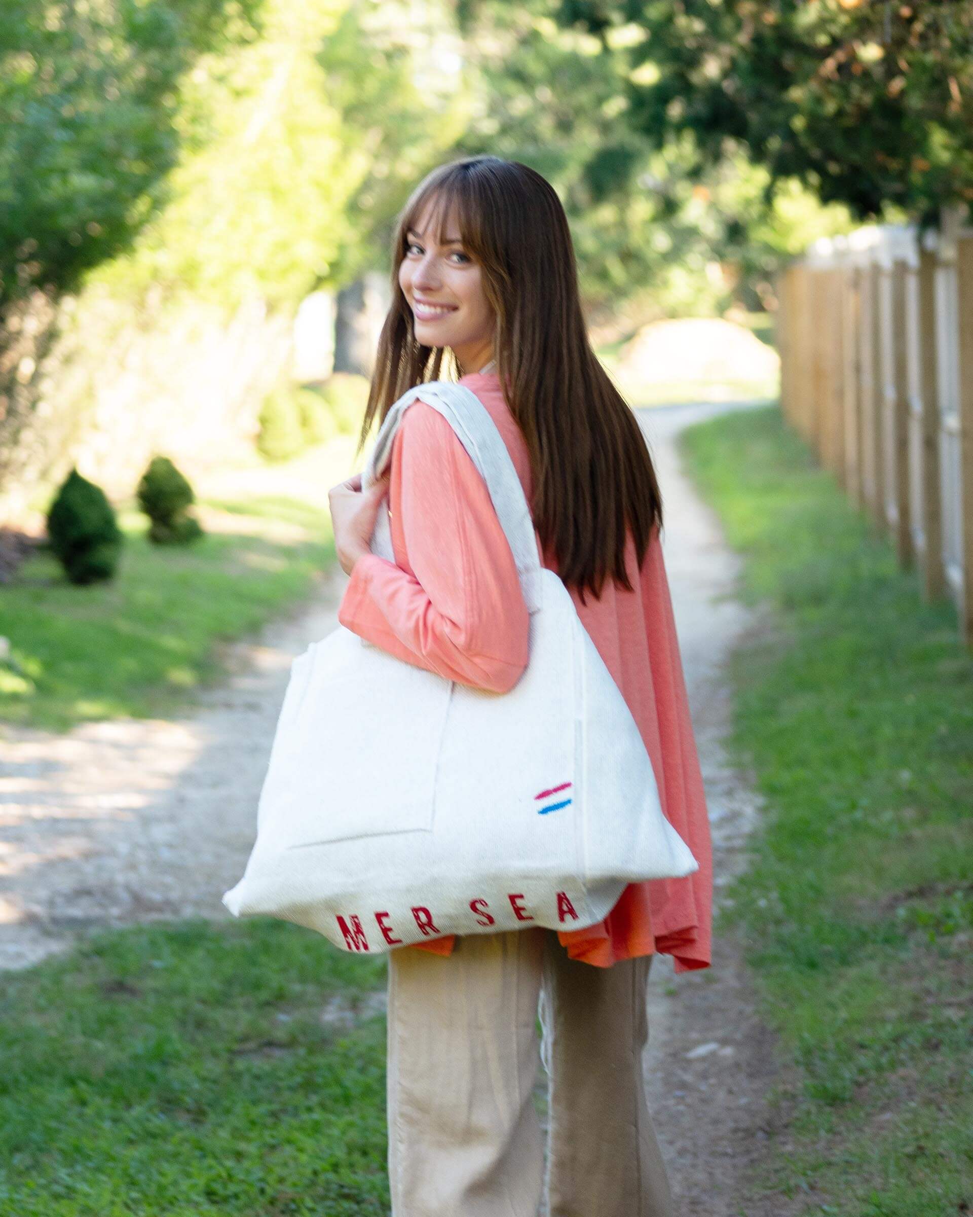 female wearing large white MERSEA labeled tote bag over one shoulder walking outside near a fence 