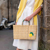 female holding medium sized straw basket with handwoven lemon leaning against a wall in the street
