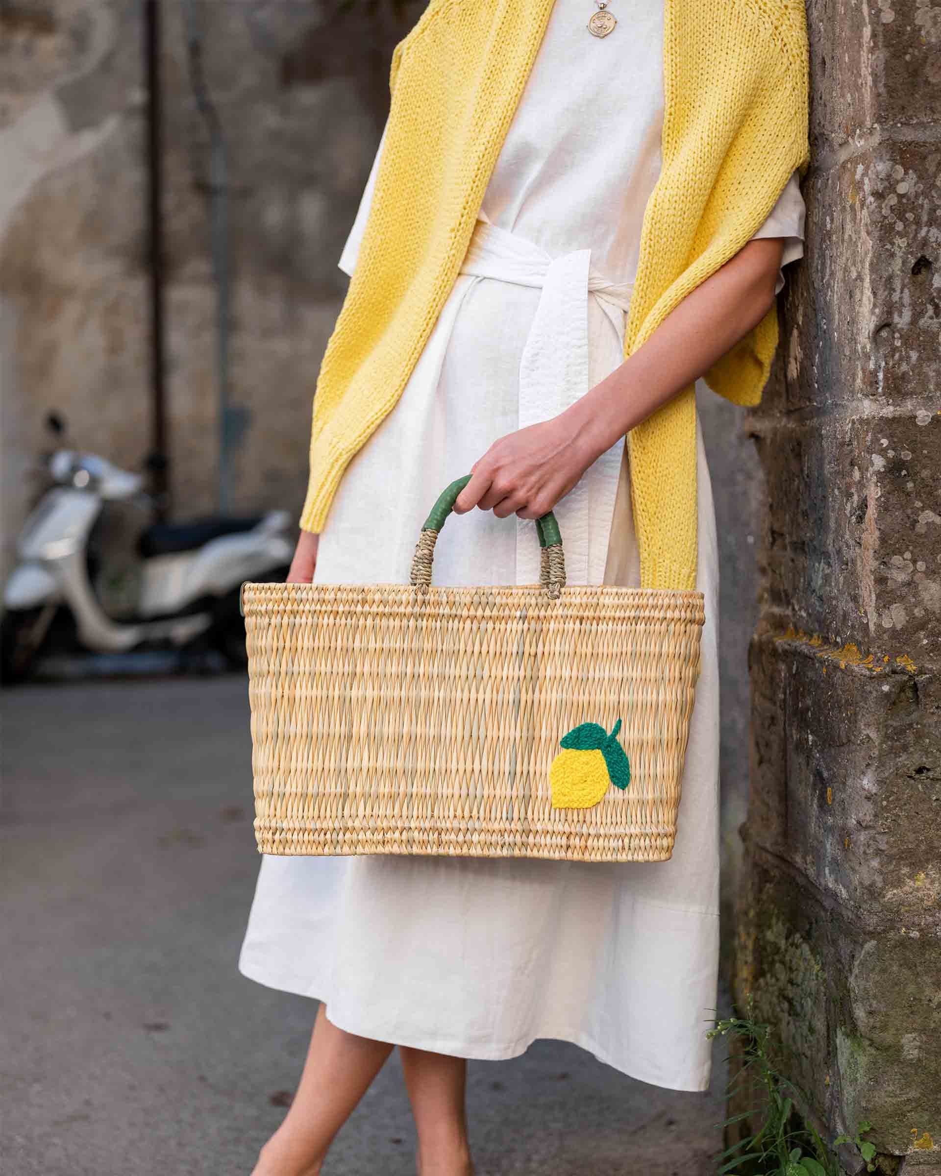 female holding medium sized straw basket with handwoven lemon leaning against a wall in the street