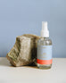 4 oz clear glass bottle of OUI! room spray next to a rock on a greyish blue background