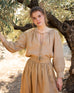 Female wearing blue jeans and beige blouse and skirt leans against an olive tree