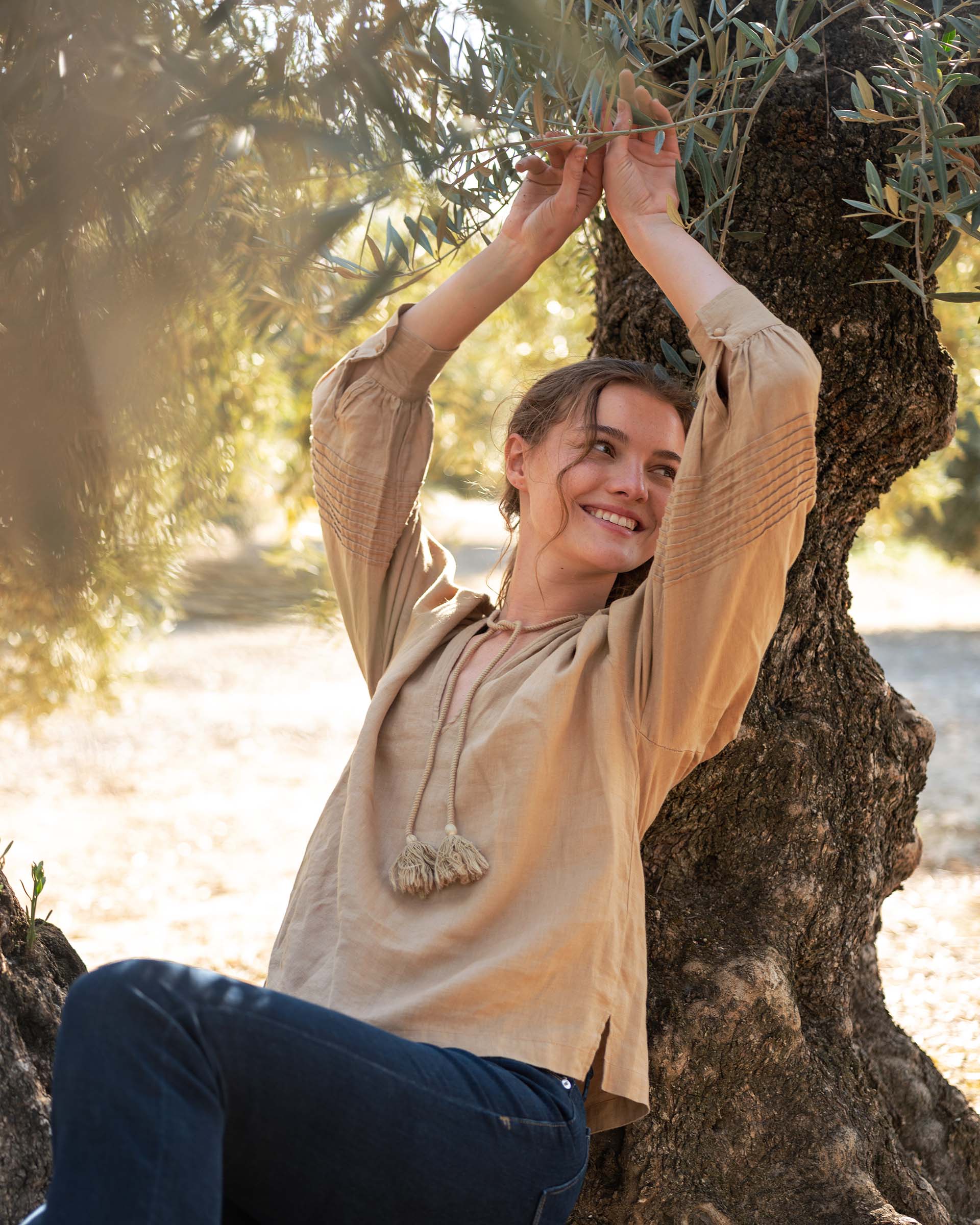 Female wearing blue jeans and beige blouse leans against an olive tree