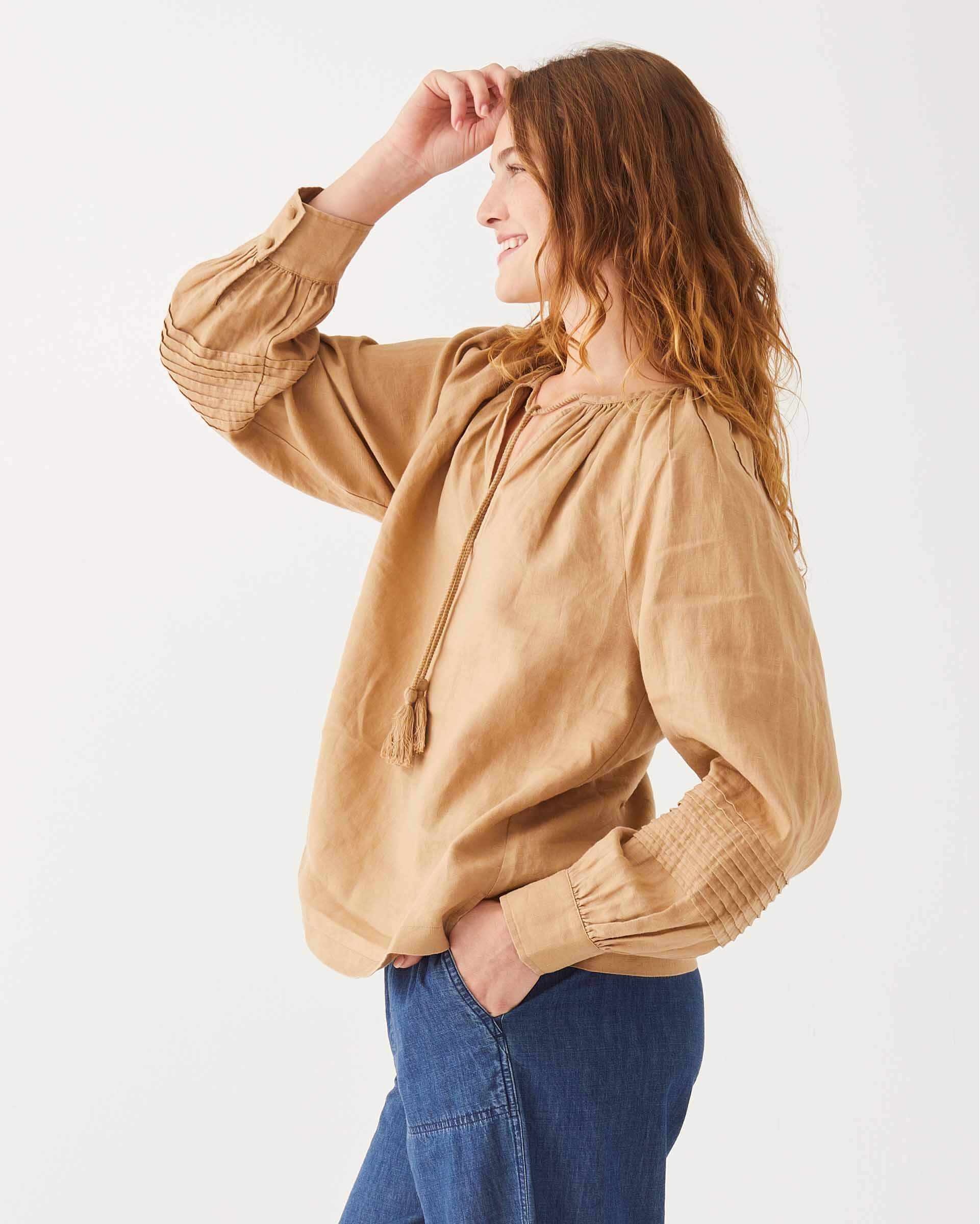 female wearing tan linen blouse with v-neck, tassels, and side slits sideways on a white background
