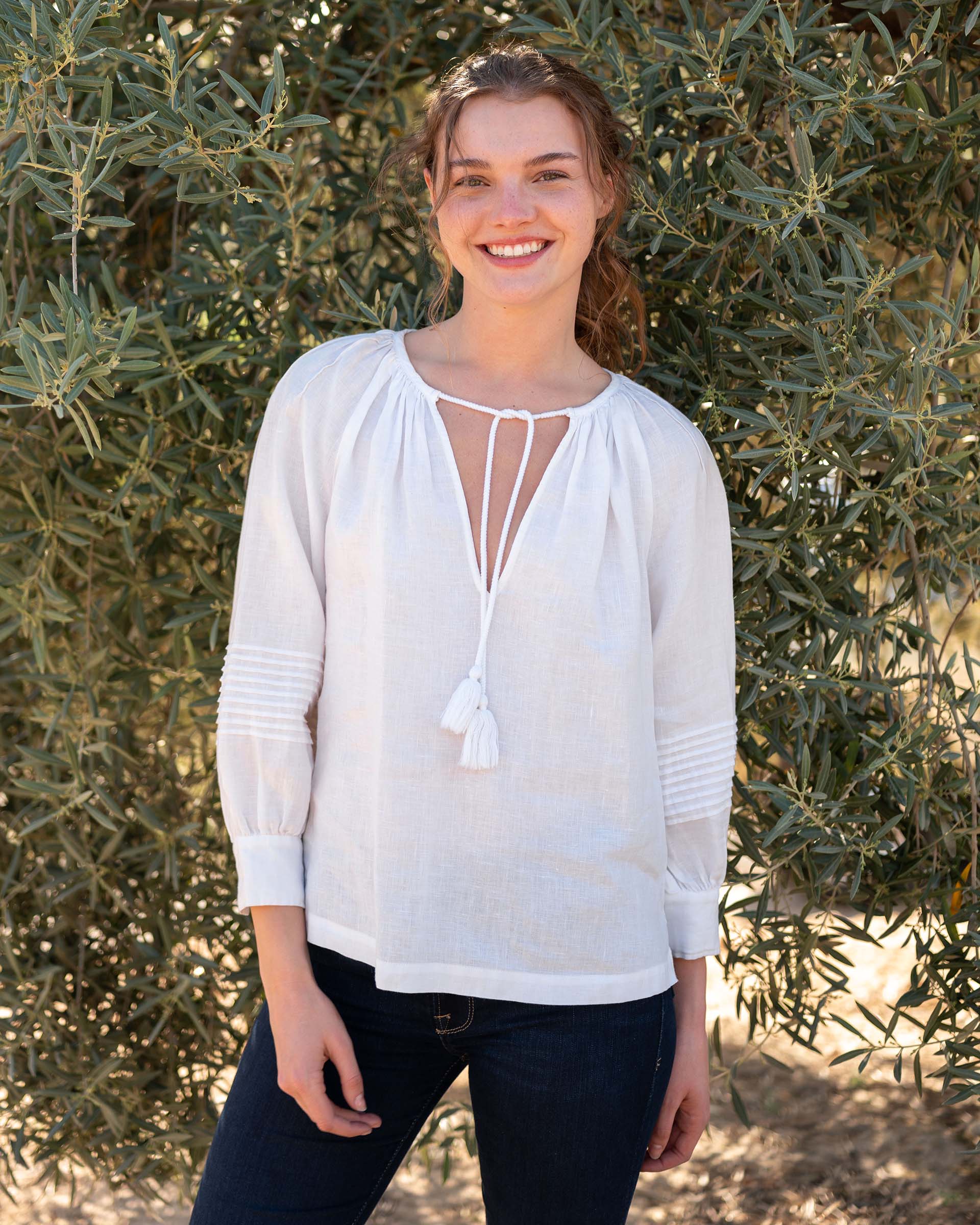 female wearing a white linen blouse and blue jeans stands smiling in front of olive tree