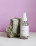 4 oz clear glass bottle of coconut sugar room spray next to a rock on a pink background