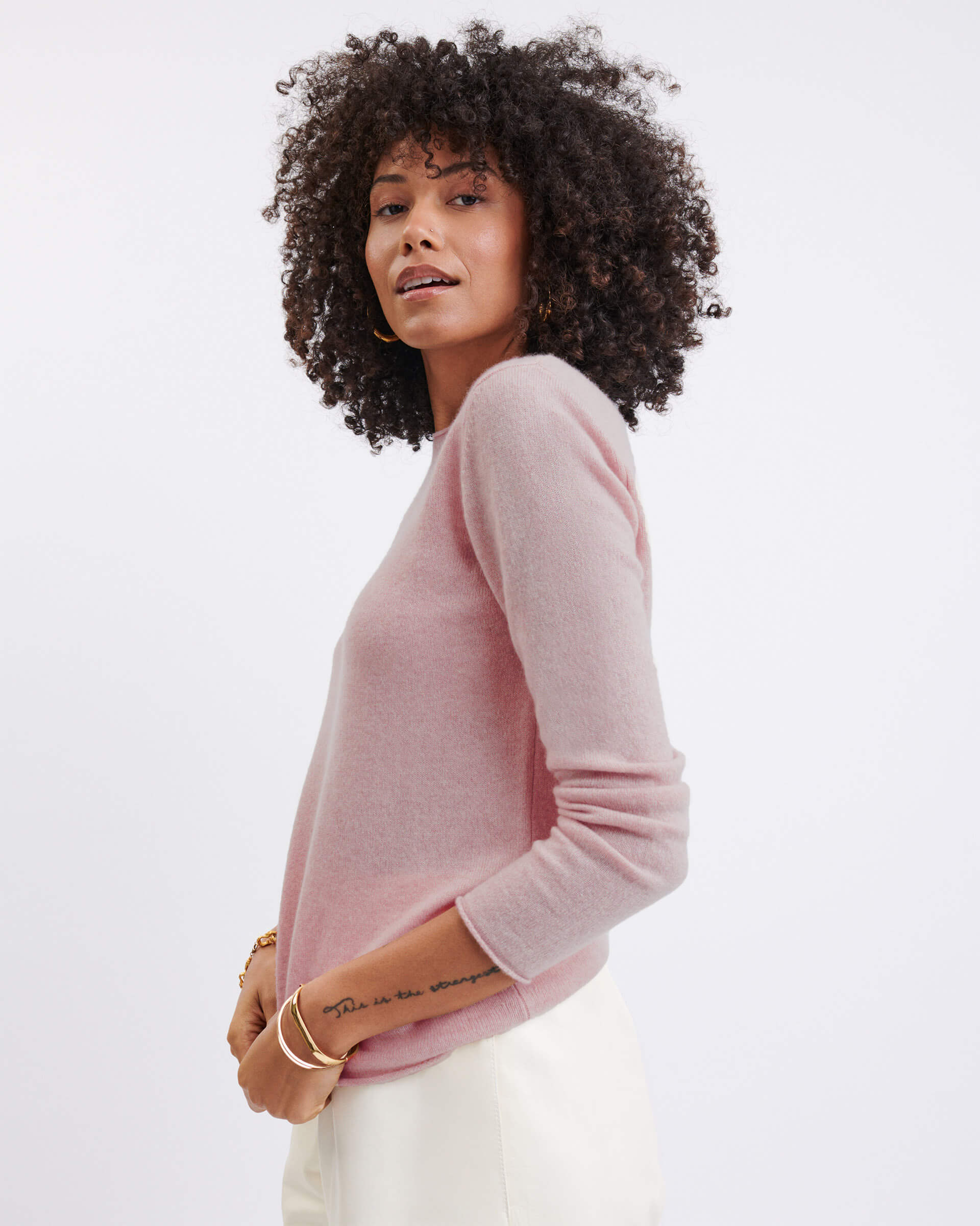 female wearing light pink sweater standing sideways holding bottom of sweater on white background 