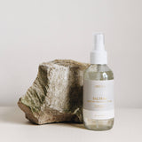 4 oz clear glass bottle of saltaire room spray next to a rock on a light grey background