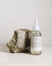 4 oz clear glass bottle of saltaire room spray next to a rock on a light grey background