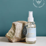 4 oz clear glass bottle of sea change room spray next to a rock on a greenish blue background