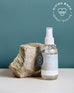 4 oz clear glass bottle of sea change room spray next to a rock on a greenish blue background