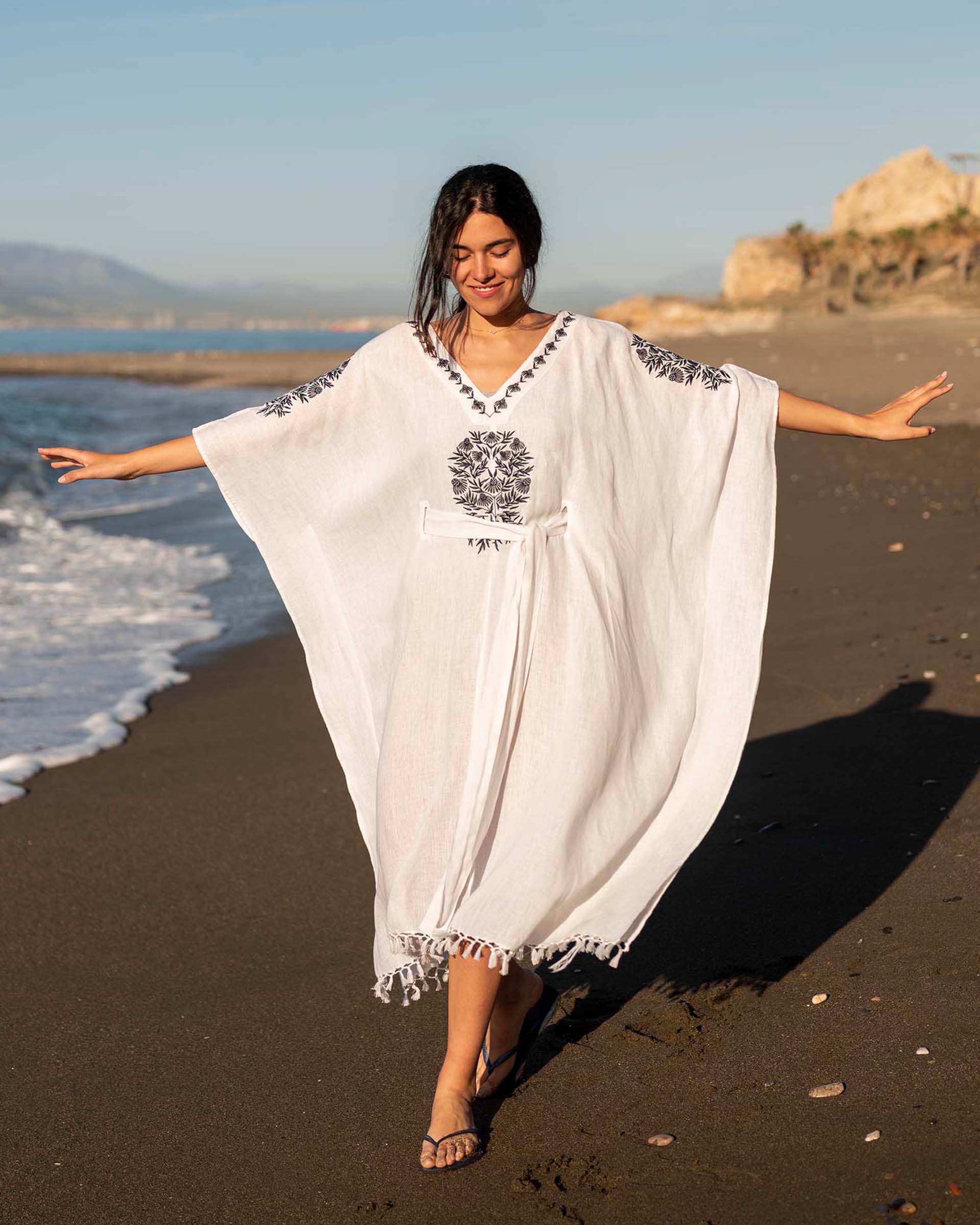 Brown hair female walks on the beach with arms wide open wearing a white cover up with embroidered details