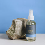 4 oz clear glass bottle of voyager room spray in dark blue label next to rock on a blue background