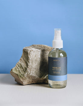 4 oz clear glass bottle of voyager room spray in dark blue label next to rock on a blue background